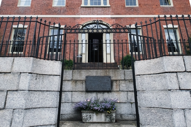 Exterior facade of a red brick mansion behind a cast iron gate around grey stone terrace.